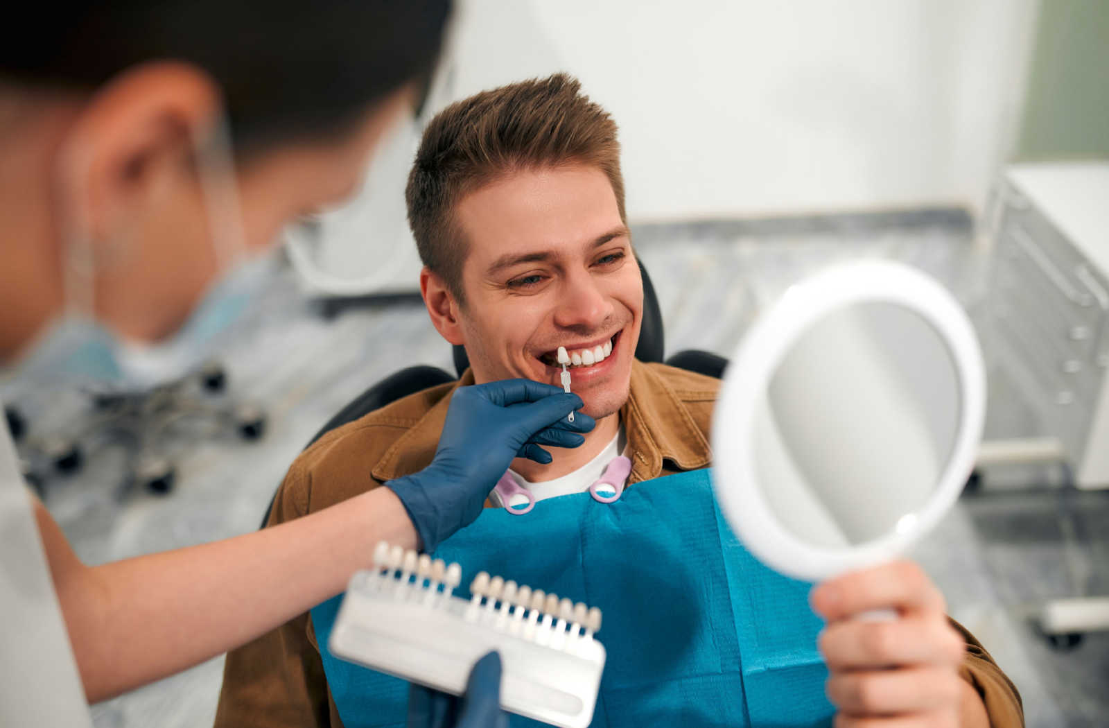 A man sitting on a dental chair is holding a mirror while the dentist is helping the patient colour match.